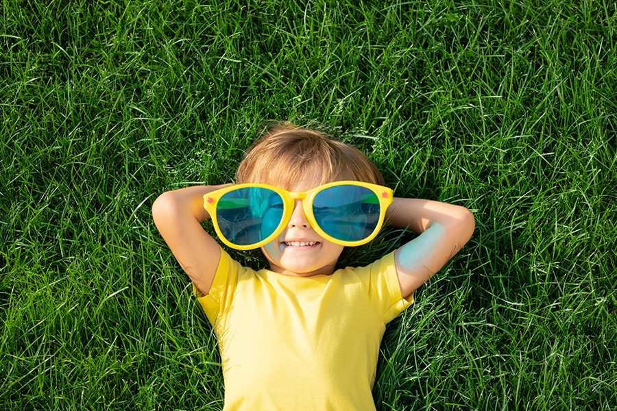 Happy child with overlarge sunglasses and a bright yellow shirt relaxing on the grass of a lush, green lawn in Springfield, IL.